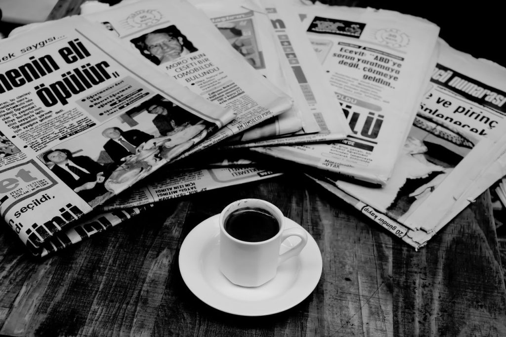 A cup of Turkish coffee sits on a wooden table next to a stack of newspapers.